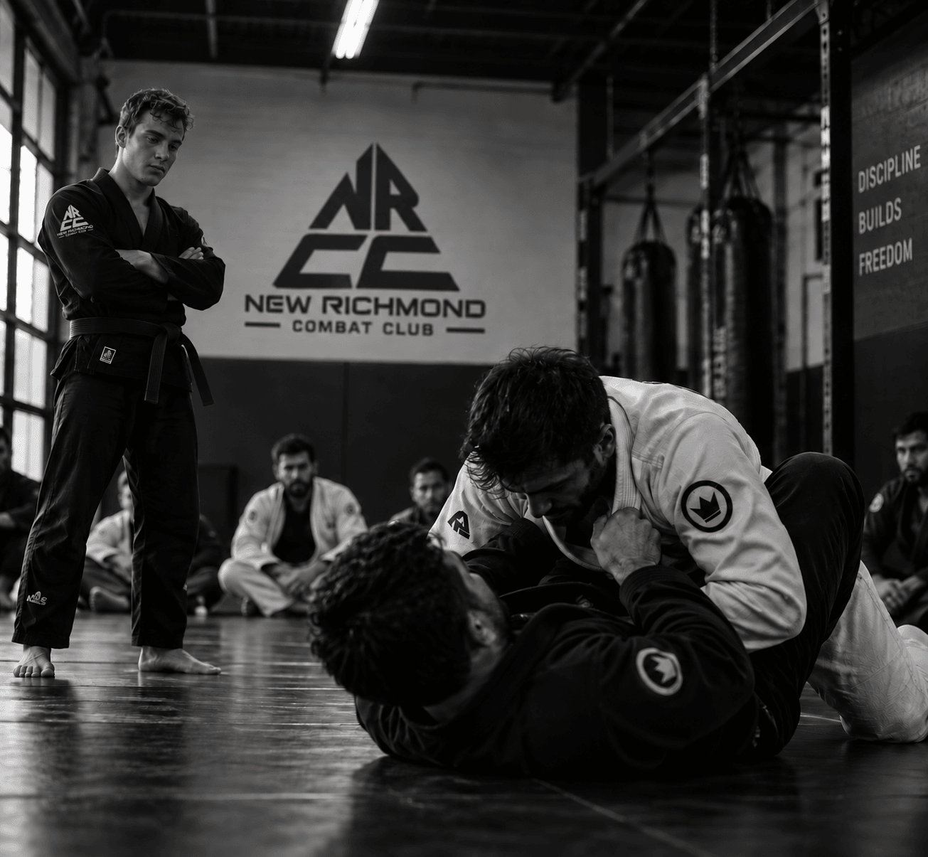Students training Brazilian jiu-jitsu on the mats, instructor watching from the side