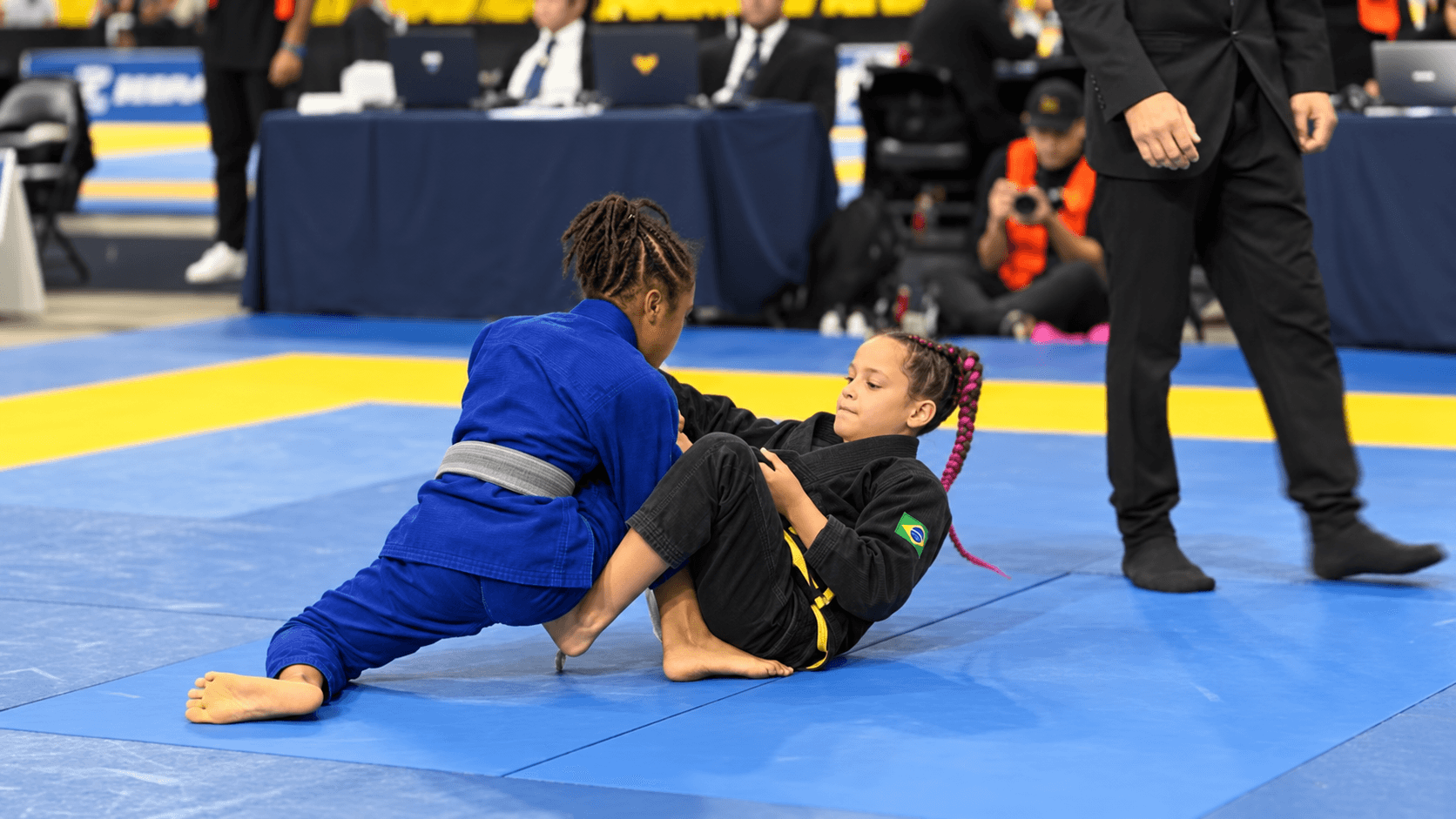 Two kids competing in jiu-jitsu — blue gi in the guard of a black gi on tournament mats